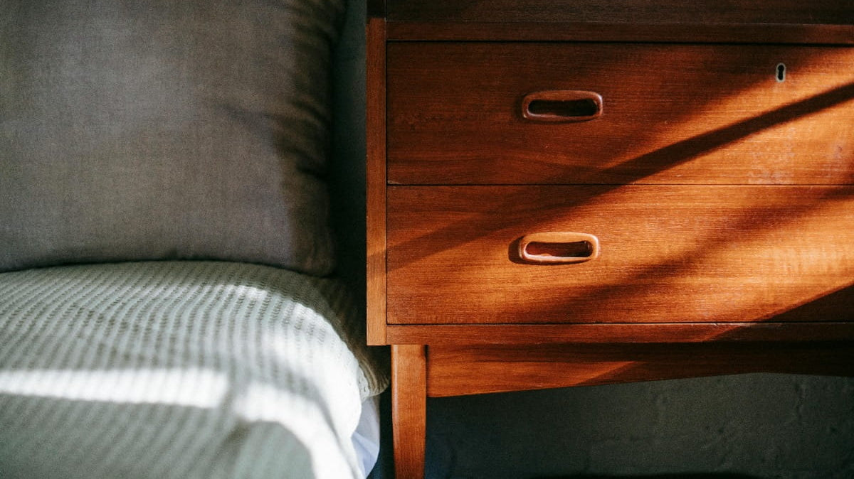 Wooden bedside table with drawers next to a bed, lit by warm sunlight casting shadows.
