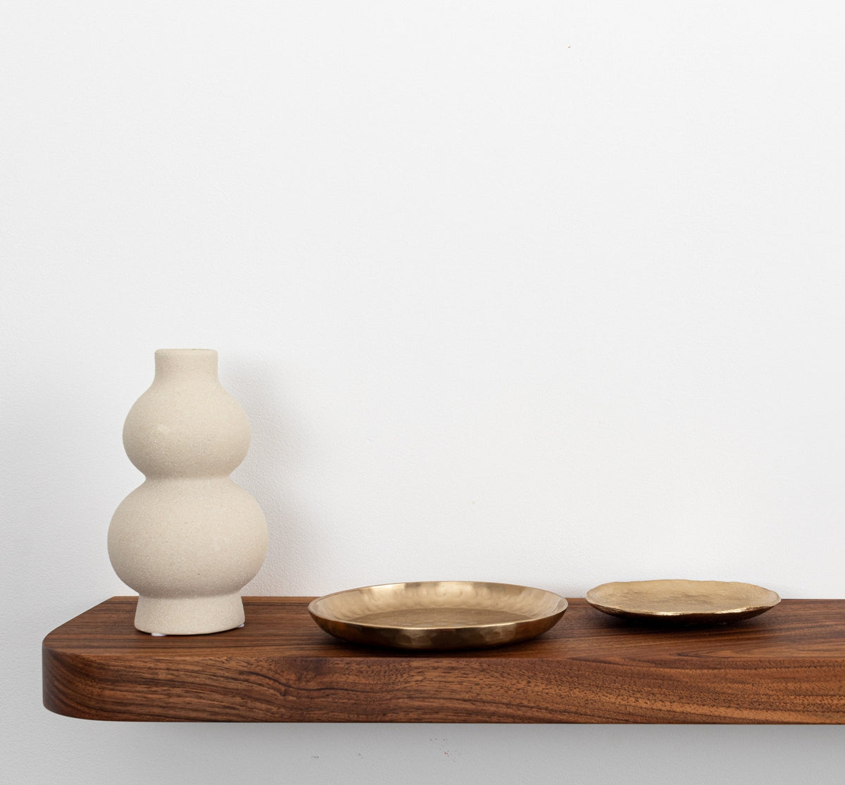 White ceramic vase and two brass plates on a wooden shelf against a white wall