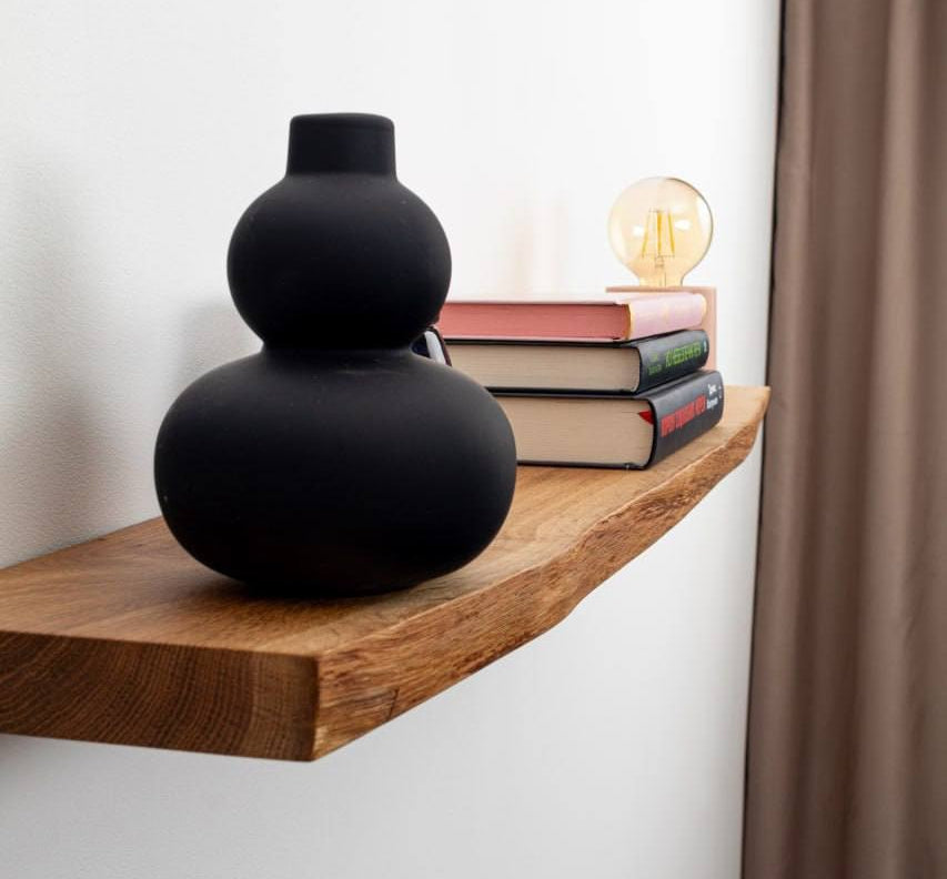 Black vase on a wooden shelf with books and a light bulb against a white wall.