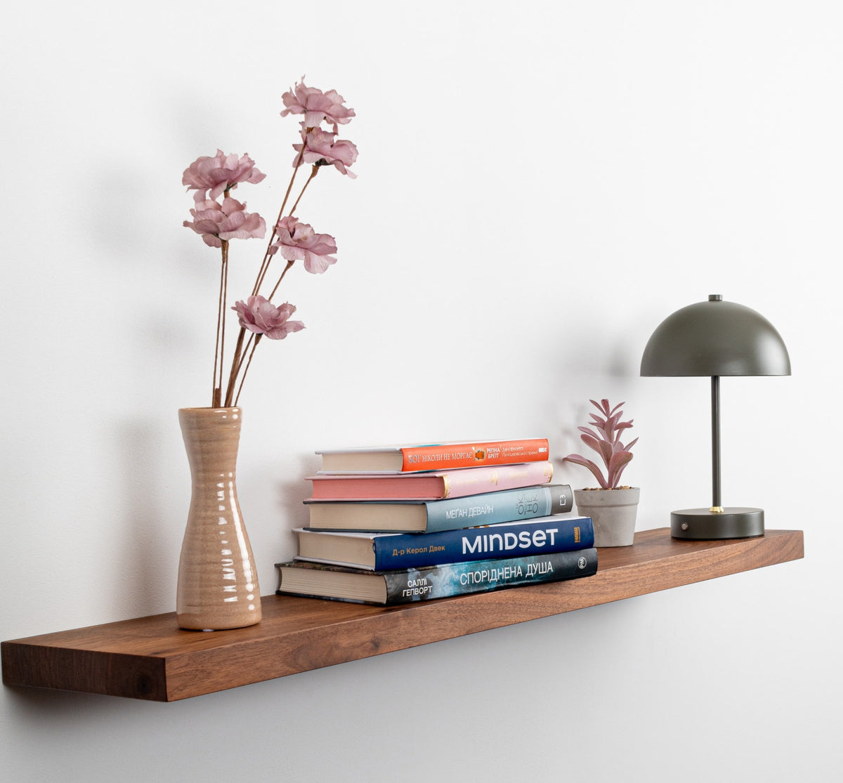 Wooden shelf with books, a vase with flowers, and a lamp against a white wall.