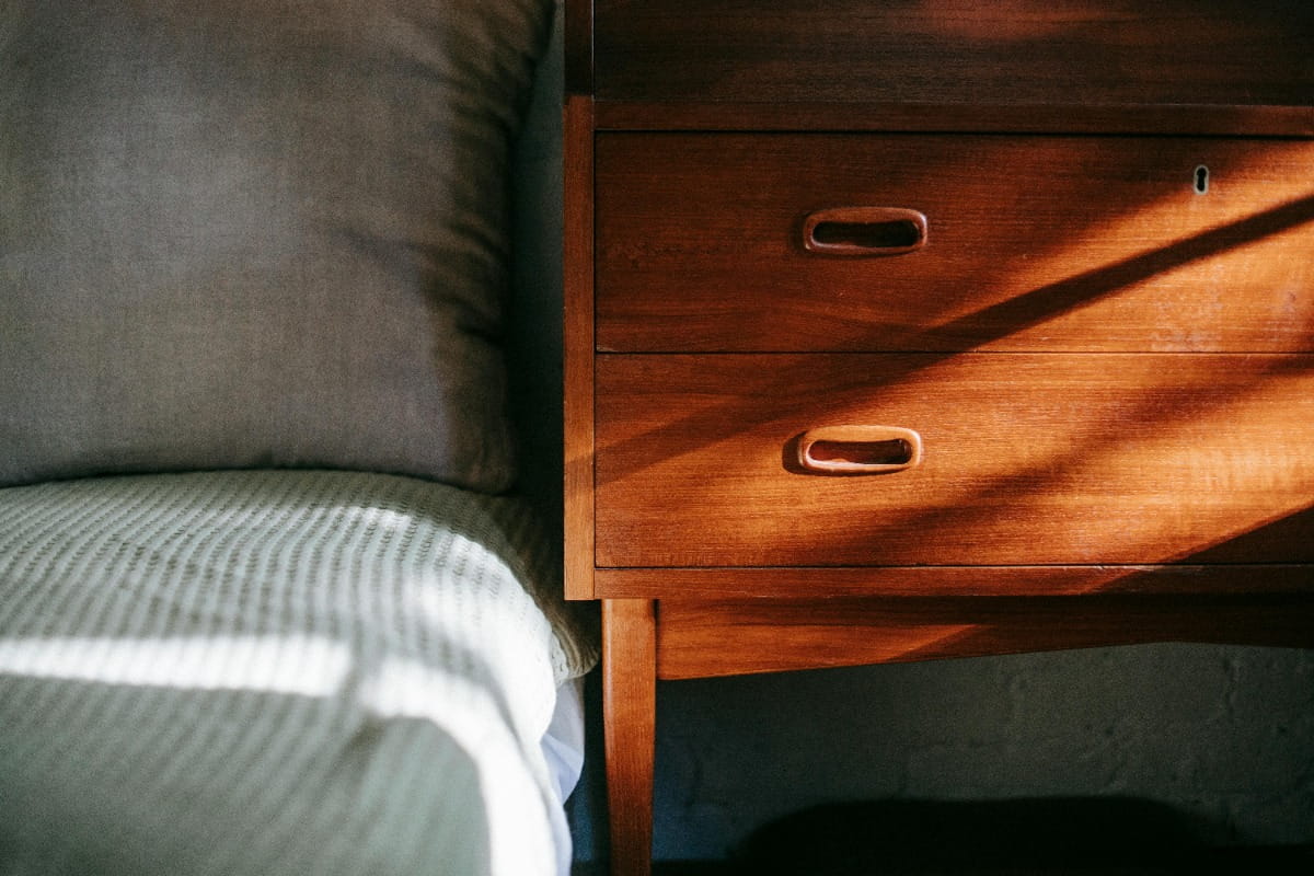 Wooden bedside table with drawers next to a bed, lit by warm sunlight casting shadows.