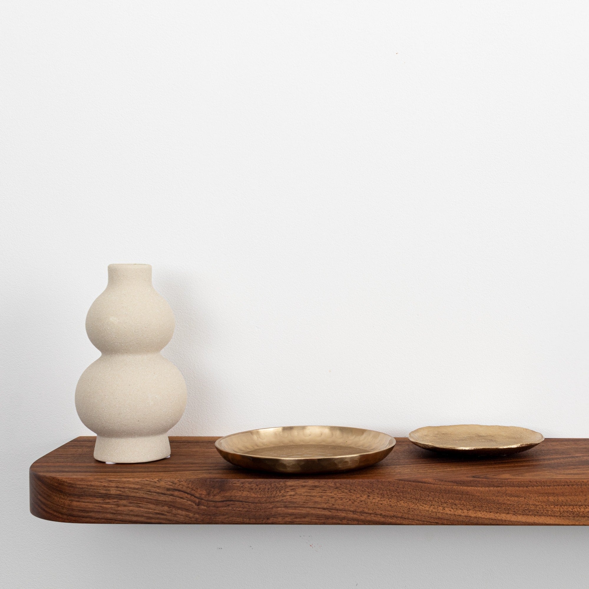 White ceramic vase and two brass plates on a wooden shelf against a white wall