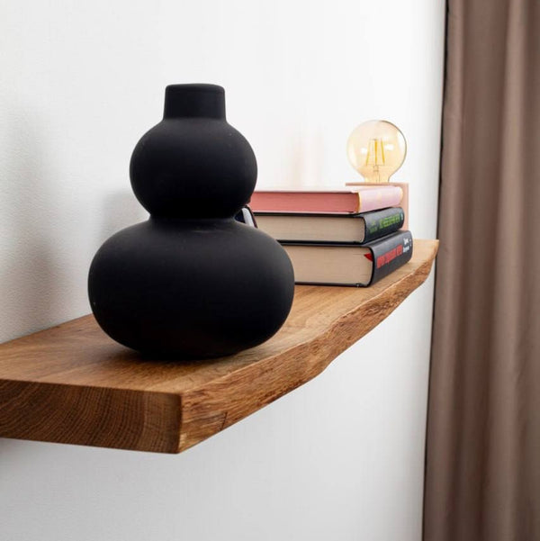Black vase on a wooden shelf with books and a light bulb against a white wall.
