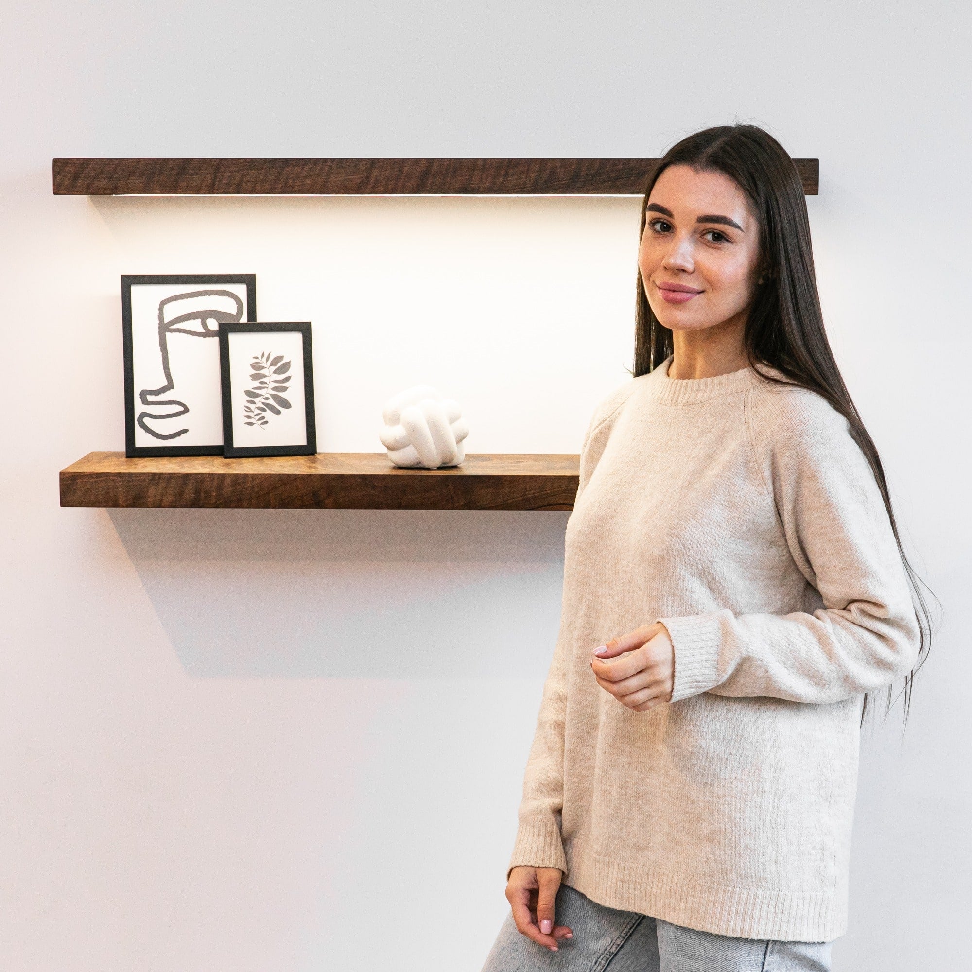 Woman in a beige sweater standing in front of a wooden shelf with decorative items.