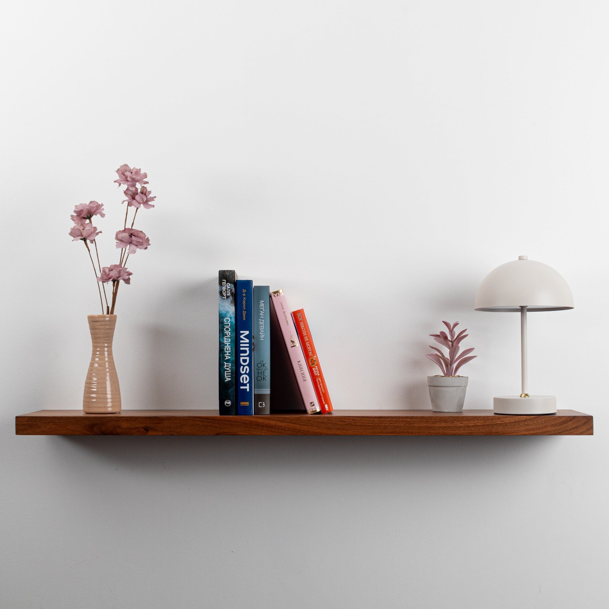 Wooden shelf with books, a vase with flowers, and a small lamp against a plain wall.