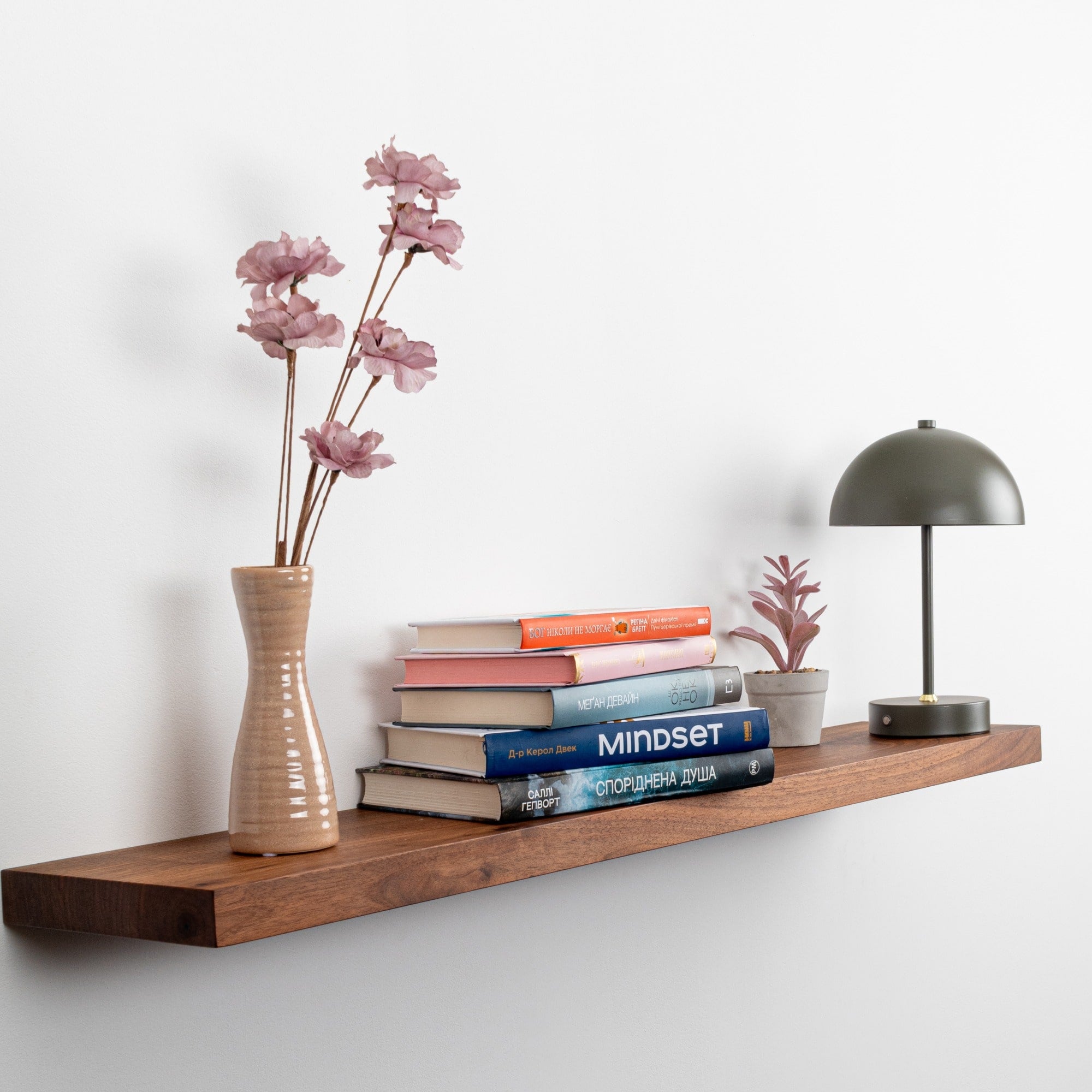 Wooden shelf with books, a vase with flowers, and a lamp against a white wall.